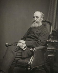 Dr William Lyon. A bearded man sits in a chair. He's not looking directly at the camera, A rolled up book is in his right hand. His left hand sits on his right