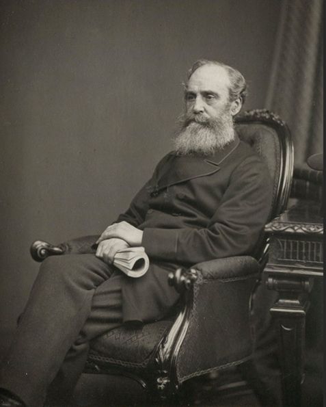 Dr William Lyon. A bearded man sits in a chair. He's not looking directly at the camera, A rolled up book is in his right hand. His left hand sits on his right