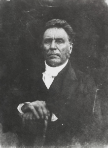 Rev. Dr. William Symington (1795-1862). Black and White image. His hands rest on a walking stick. He wears a Minister's robes